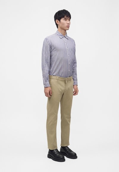 Jeune homme debout portant une chemise à boutons rayée bleu et blanc, un pantalon beige et des mocassins noirs sur un fond blanc uni.