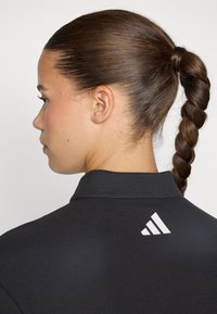 Black polo shirt with a textured fabric, featuring a white Adidas logo at the back near the collar. Hair styled in a braid.