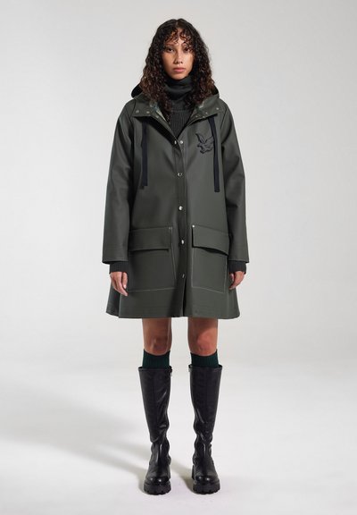 Abrigo verde impermeable con capucha, que cuenta con bolsillos frontales, un logo bordado en negro y un corte relajado. Botas de goma negras altas completan el atuendo.