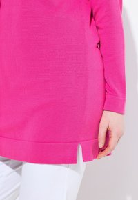 Túnica fucsia de manga larga con textura acanalada, con aberturas en los laterales del dobladillo, combinada con pantalones blancos recortados.