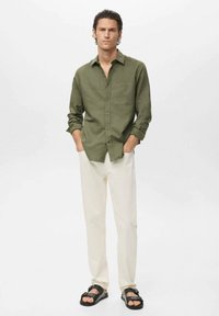 Camisa de botón verde oliva con un bolsillo en el pecho, combinada con pantalones color crema y sandalias negras. Textura suave, corte relajado.