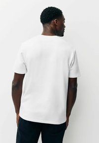 Fehér, rövid ujjú póló kerek nyakkivágással, sima felülettel és laza szabással, hátulnézetből, semleges háttér előtt.