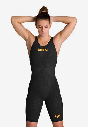 Vrouwelijke zwemmer in een zwart badpak met gele "arena"-logo, staand met haar handen achter haar hoofd tegen een witte achtergrond.