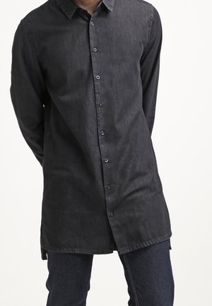 Mand bærer en lang sort skjorte med knapper og krave samt mørkeblå jeans, står med armene bag ryggen mod en hvid baggrund.