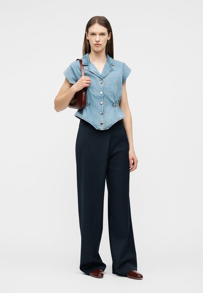 Femme portant un haut en jean sans manches à boutons, un pantalon noir à jambes larges, des chaussures marron, et un petit sac à bandoulière marron.