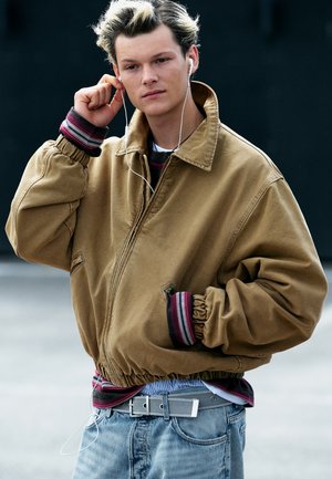 Jeune personne portant une veste marron et un jean ajuste ses écouteurs d'une main, debout devant un fond sombre uni.