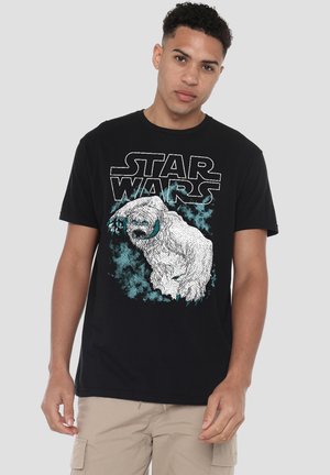 Junger Mann trägt ein schwarzes Star Wars T-Shirt mit weißem und türkisfarbenem Monster-Design, steht vor einem schlichten hellgrauen Hintergrund.