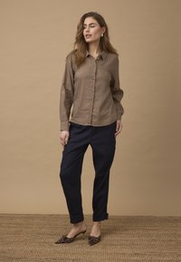 Femme debout contre un mur beige portant une chemise marron boutonnée, un pantalon noir, des talons pointus à imprimé léopard et des boucles d'oreilles dorées.
