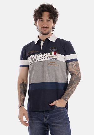 Uomo con capelli ricci che indossa una polo blu navy, grigia e bianca con loghi Aeronautica Militare e tatuaggi su entrambe le braccia, posando con una mano in tasca.
