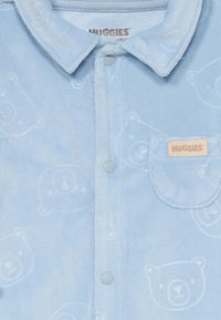 Hellblauer Baumwoll-Body mit einem flauschigen Material, Knopfverschluss und einem geprägten Bärenmuster. Enthält eine kleine Tasche mit einem Etikett.