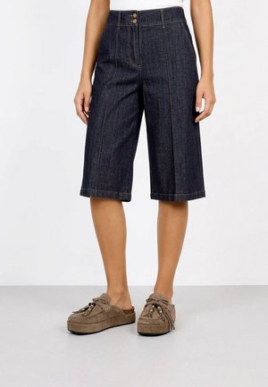 Mujer con culottes de mezclilla oscura hasta la rodilla y mocasines taupe con detalles de borlas, de pie sobre fondo blanco.