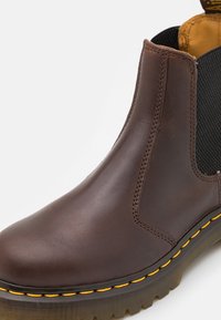 Bottine en cuir brun avec panneaux élastiques noirs sur les côtés et coutures jaunes. Dispose d'une semelle en caoutchouc texturée pour une meilleure adhérence.