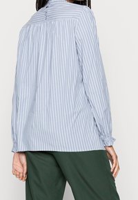 Blouse rayée bleu clair avec manches longues, détail froncé à l'arrière, et tissu doux. Associée à un pantalon vert foncé.