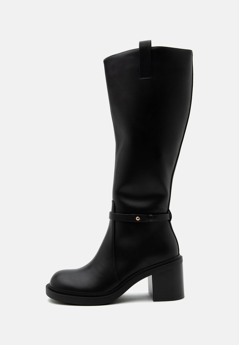 Botte en cuir noir montant jusqu'au genou avec un talon bloc et un détail de sangle à la cheville sur fond clair uni.