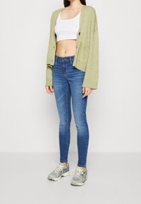 Cárdigan oversized verde claro, top corto blanco, jeans ajustados azules y zapatillas grises. El cárdigan presenta botones frontales y bolsillos.