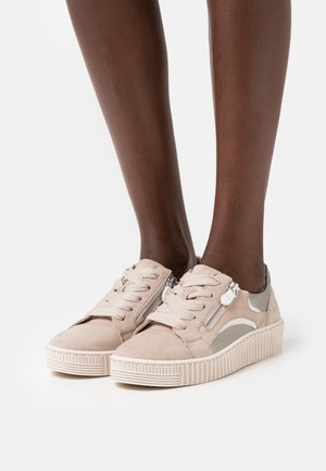Baskets compensées en daim beige avec lacets et fermetures éclair latérales portées sur des pieds à peau foncée sur fond blanc.