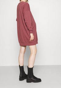 Abito felpa rosso porpora oversize con maniche lunghe e scollatura tonda, abbinato a stivaletti neri alla caviglia con pannelli elastici laterali.