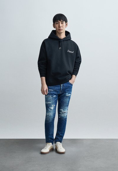 Μαύρο φούτερ με κουκούλα και κορδόνι, με λευκό κείμενο. Φορεμένο με σκισμένο μπλε τζιν και κρεμ αθλητικά παπούτσια με χρυσές λεπτομέρειες.