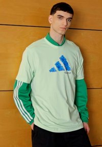 Maglietta a maniche corte verde chiaro con il logo Adidas testurizzato blu. Indossata sopra una maglietta a maniche lunghe verde, con strisce bianche sulle maniche.