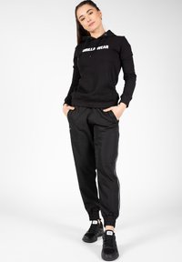 Zwarte hoodie met "GORILLA WEAR" in het wit gedrukt, gecombineerd met zwarte joggers met een slim fit en witte zijkanten, vergezeld van zwarte sneakers.