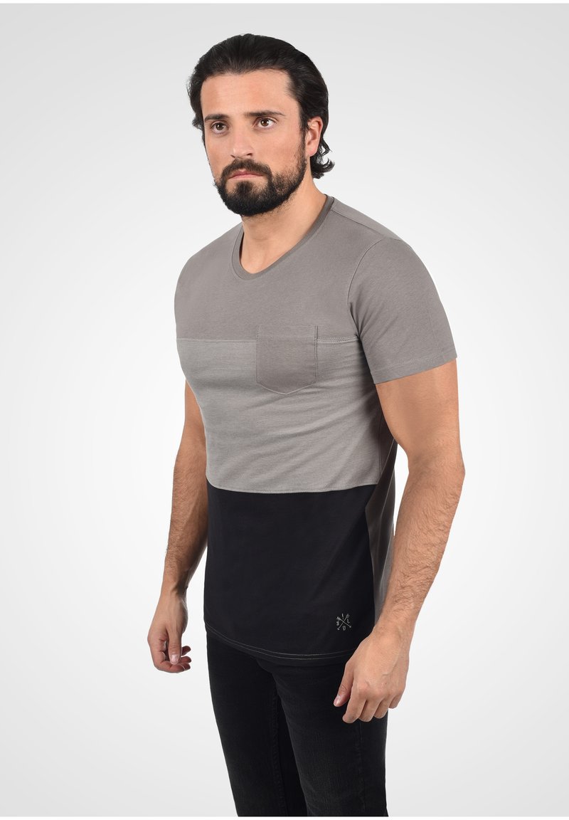 Hombre con cabello y barba oscura, vistiendo una camiseta de color bloque gris y negro de mangas cortas y pantalones negros, de pie contra un fondo liso.