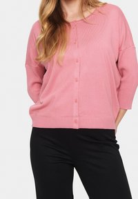 Pink cardigan with a button front, ribbed cuffs, and hem. It has a relaxed fit and is worn over black pants, displaying a soft texture.