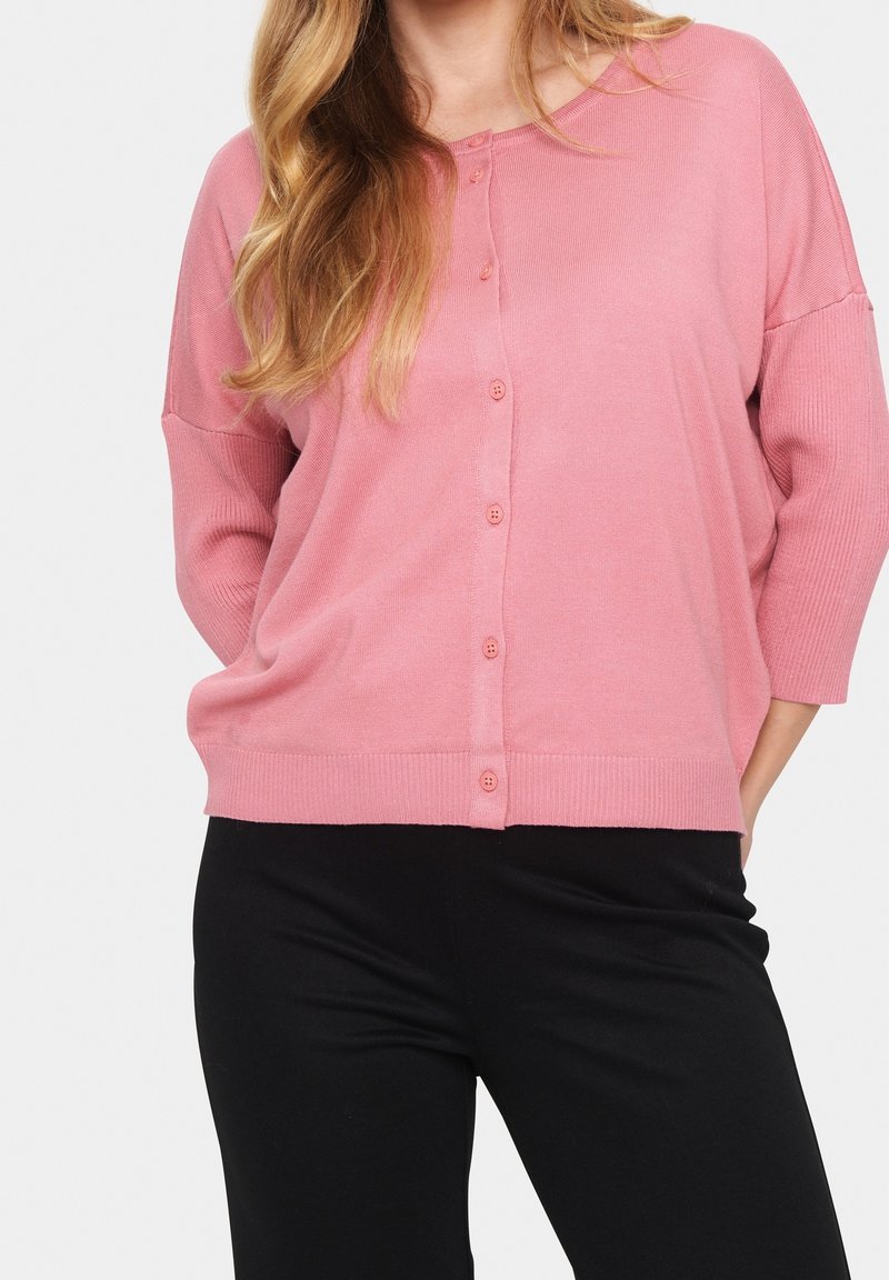 Pink cardigan with a button front, ribbed cuffs, and hem. It has a relaxed fit and is worn over black pants, displaying a soft texture.