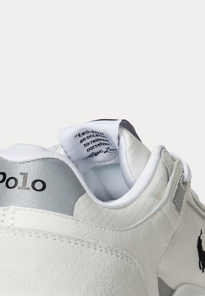Baskets blanches Polo montrant le talon arrière, l’étiquette intérieure de la languette avec texte et le logo Polo brodé noir sur le côté.