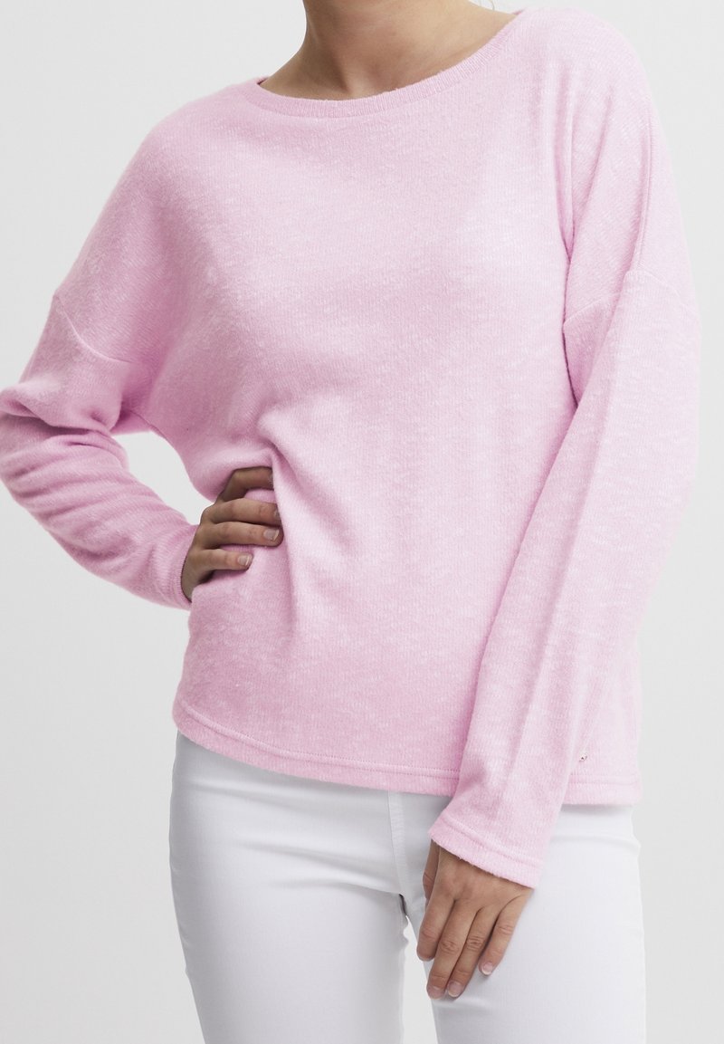Personne portant un pull rose clair à manches longues et un pantalon blanc, debout avec une main sur la hanche et l'autre détendue.