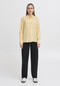 Camicia a righe gialle con colletto, dotata di maniche lunghe. Abbinata a pantaloni neri e sneakers bianche, creando un look casual.