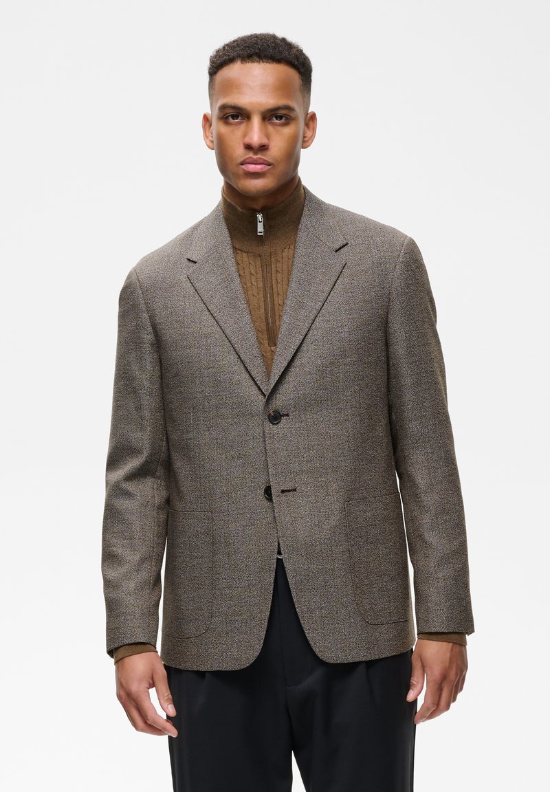 Homme portant un blazer en tweed marron sur un pull zippé marron et un pantalon noir, debout devant un fond blanc uni.