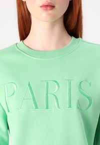 Svetlo zelena majica s crewneck izrezom, ki ima izbočeno vezenje besede "PARIS" v usklajeni barvi preko prsi. Gladka tekstura.