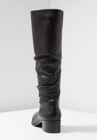 Bota alta de cuero negro con un diseño arrugado, que cuenta con un robusto tacón de bloque y una textura suave, contra un fondo neutro.