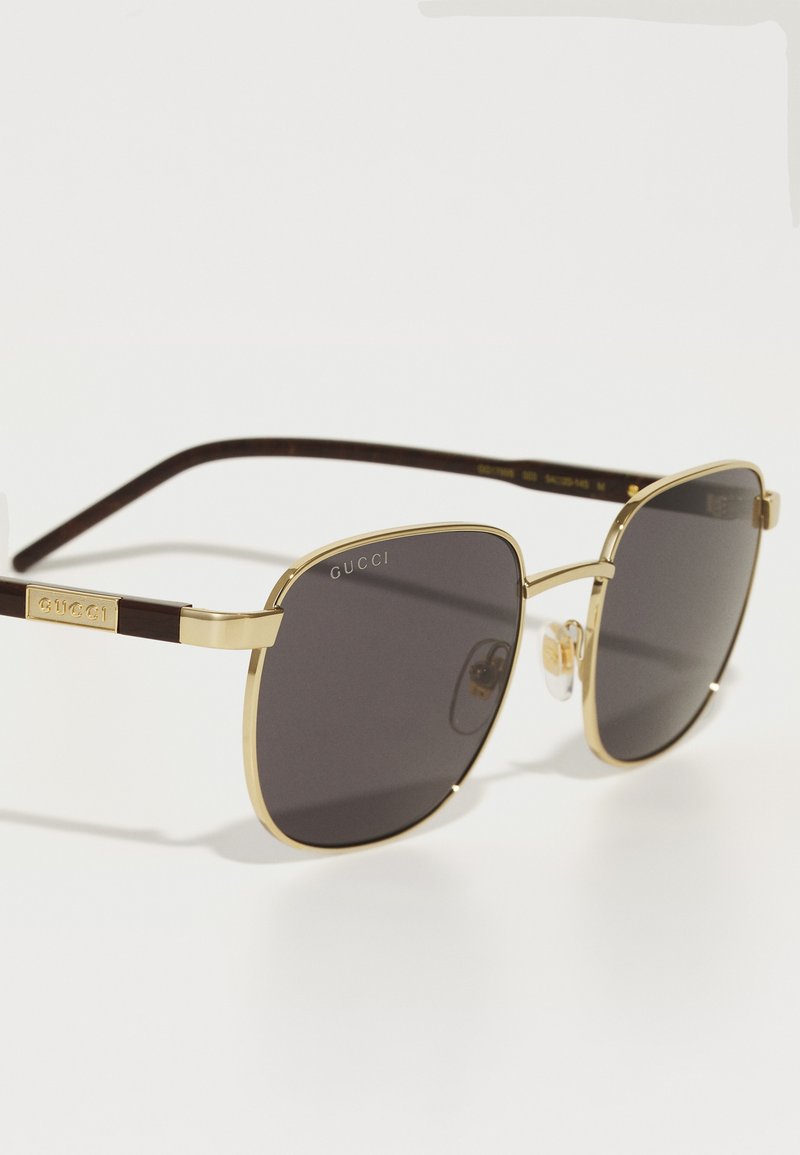 Lunettes de soleil Gucci avec monture en métal doré et verres gris foncé. Branches en écaille de tortue marron avec détail de logo et plaquettes de nez transparentes. Forme carrée.