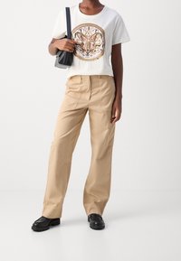 Bílý bavlněný tričko s grafickým motivem motýla, béžové široké kalhoty, černá kabelka přes rameno a masivní černé boty. Hladký materiál a volný střih.