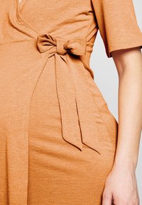 Sukienka w kolorze rdzy z dekoltem w serek, krótkimi rękawami i bocznym węzłem, który tworzy marszczony efekt. Gładki, miękki materiał.