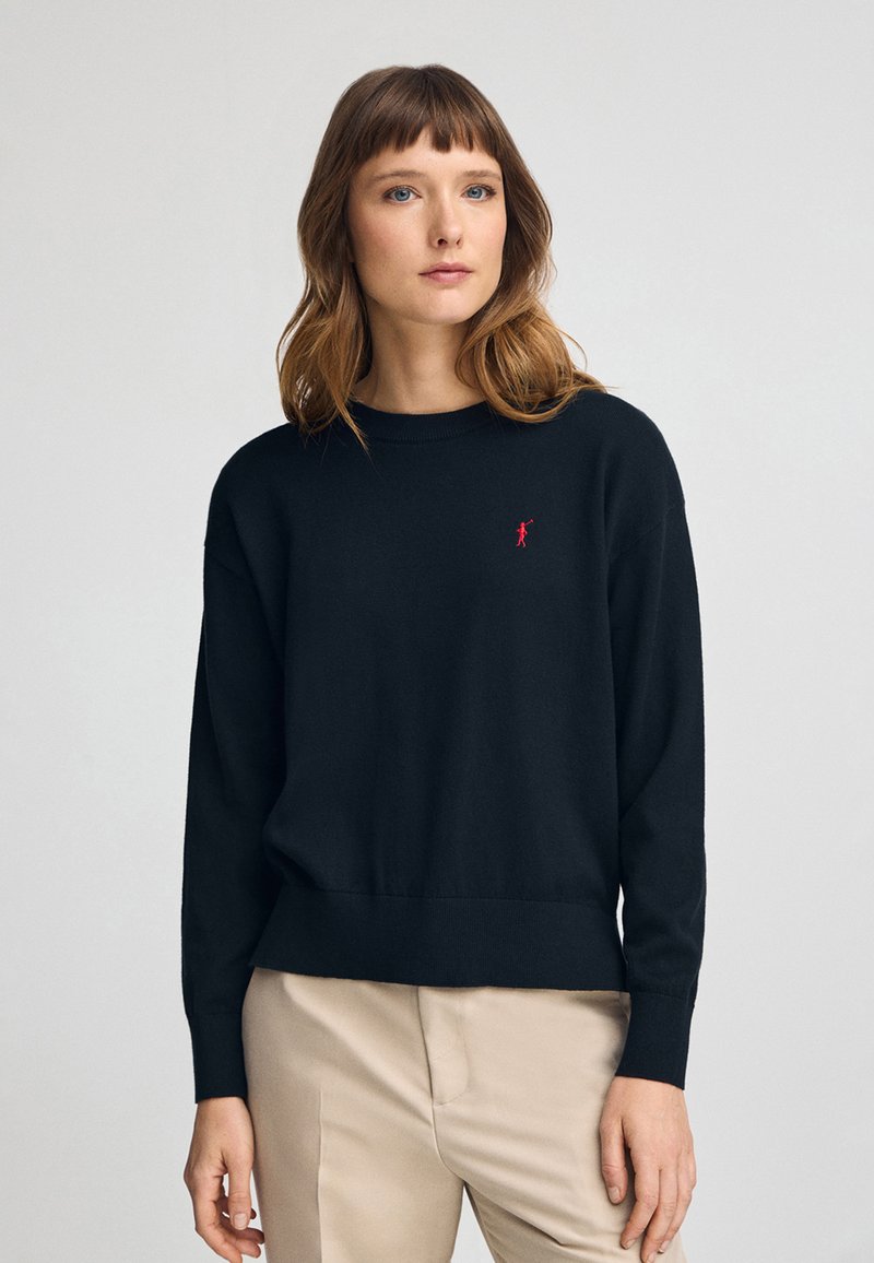 Suéter negro de punto con cuello redondo, mangas largas, dobladillo acanalado y pequeño logo rojo en el pecho. Textura suave, ajuste casual.