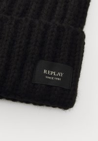 Sort strikket hue med ribbet tekstur. Har et rektangulært mærkat med "REPLAY" og "SINCE 1981" i hvid tekst. Blød, varm og strækbar.