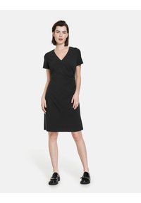 Schwarzes Kurzarmkleid mit V-Ausschnitt, figurbetontem Schnitt, glatter Textur und Seitenschlitzen, kombiniert mit schwarzen flachen Schuhen.