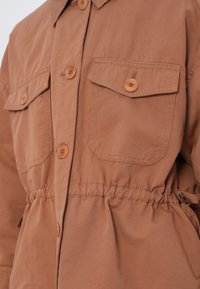 Veste marron boutonnée avec deux poches avant à rabat et un détail de taille élastique.