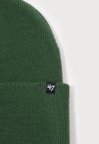 Zielona wełniana czapka beanie, wykonana z miękkiego materiału, z podwiniętym mankietem i małą czarną metką z białym tekstem z boku.