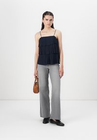 Haut plissé bleu marine avec trois niveaux, associé à un jean large gris. Le mannequin tient un petit sac à main marron et porte des chaussures noires.