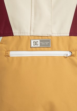 DC-jack met een bordeauxrode en crèmekleurige bovenkant en een gele ondersectie. Bevat een horizontale witte ritszak en een merklabel.