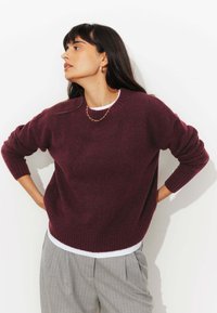 Pull bordeaux en tricot doux avec des poignets et un ourlet côtelés, superposé à une chemise blanche. Pantalons à rayures gris visibles à la taille.