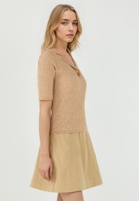 Top en tricot côtelé beige avec des manches courtes et un petit col, orné d'un accent doré. Associé à une jupe évasée de couleur beige clair.