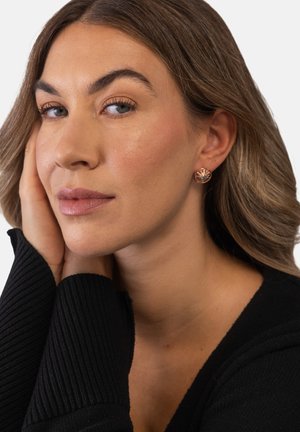 Frau mit hellbraunen Haaren, die runde roségoldene Ohrringe und einen schwarzen Pullover trägt und ihr Gesicht auf ihre Hand stützt, vor einem schlichten Hintergrund.