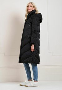 Doudoune noire avec capuche, coupe longue, motif matelassé en chevron, avec poches latérales et fermeture éclair. Portée avec un jean bleu et des baskets blanches.