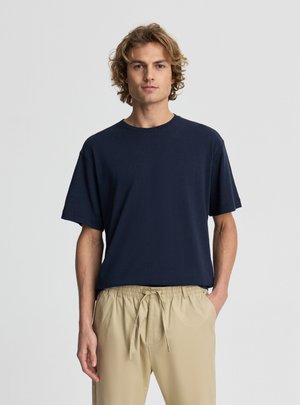 Giovane uomo con capelli biondi ricci che indossa una maglietta blu navy a maniche corte e pantaloni beige con coulisse, in piedi con una mano in tasca.