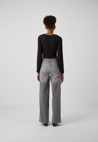 Femme debout, dos à la caméra, portant un haut noir à manches longues, un jean gris à jambes larges et des chaussures noires cloutées sur un fond uni.