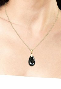 Collana in oro con un ciondolo in pietra nera a forma di pera con superficie sfaccettata, appoggiata su pelle nuda.